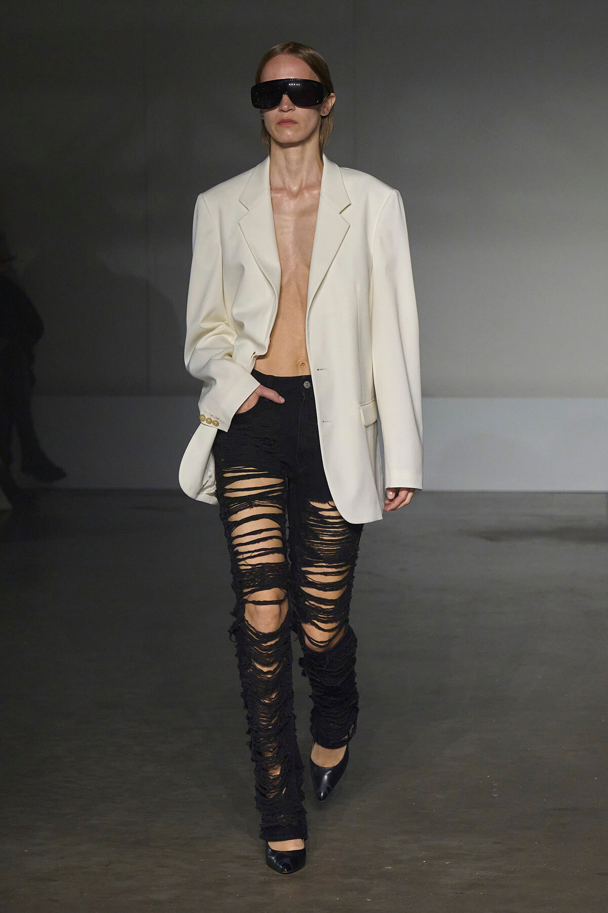 Model walking a runway in an oversized cream blazer, extremely ripped black jeans, black pointed heels, and oversized dark sunglasses.