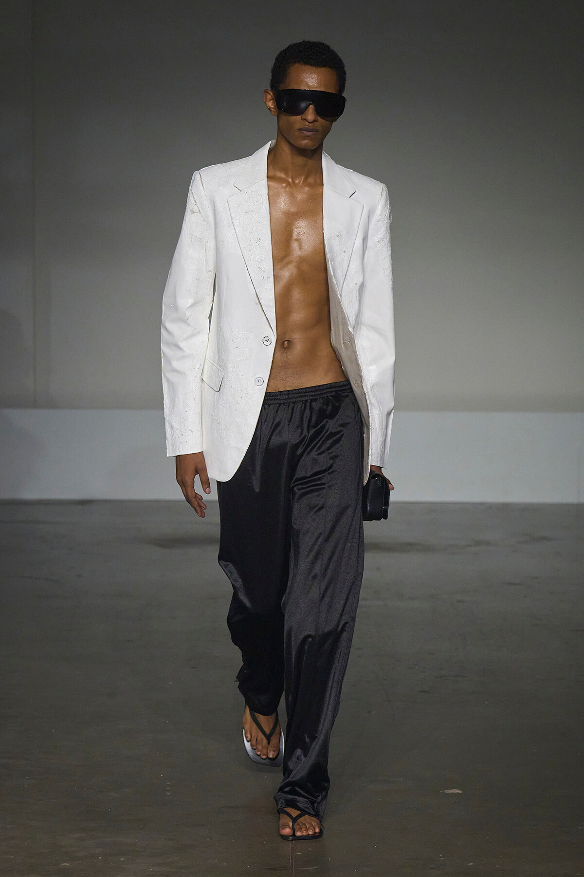 Male model on a runway wearing an open white blazer, black satin pants, oversized sunglasses, and flip-flop sandals.