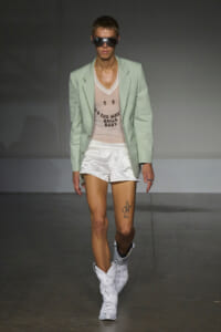 Model walks the runway in a mint blazer over a pale pink slogan tee, white satin shorts, and marble-pattern cowboy boots, with oversized sunglasses.