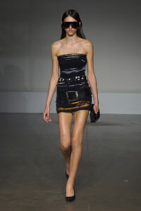 Model on a runway wearing a strapless black mini dress with a wide belt, black heels, and oversized dark sunglasses while holding a small clutch.