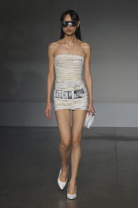 Model walking the runway in a cream ruched strapless mini dress with a wide belt, white heels, and oversized sunglasses.
