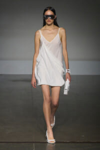 Model walks the runway in a white satin slip dress with thin straps, oversized sunglasses, white sandals, and a small white clutch.