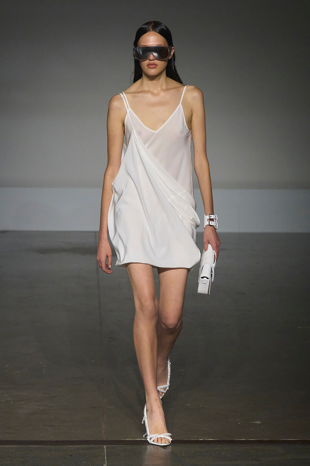 Model walks the runway in a white satin slip dress with thin straps, oversized sunglasses, white sandals, and a small white clutch.