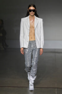 Model on runway wearing a white blazer over bare torso, glittery silver pants, and white boots with oversized sunglasses.