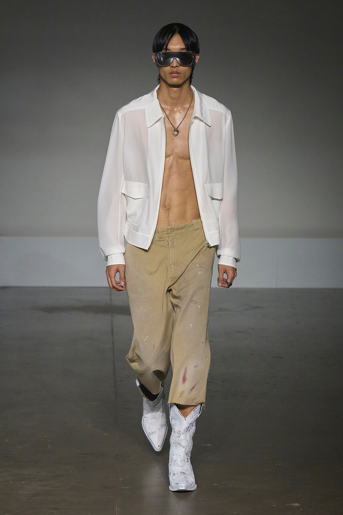 Model walks the runway in a sheer white jacket, no shirt, beige paint-splattered pants, and white boots, with oversized sunglasses and a circular pendant necklace.
