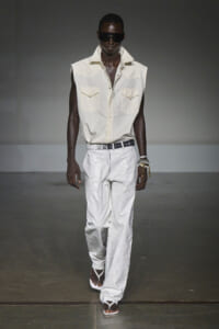 Male model on a runway wearing a sleeveless beige shirt with two breast pockets, white paint-splattered pants, a white belt, dark sunglasses, and white sandals.