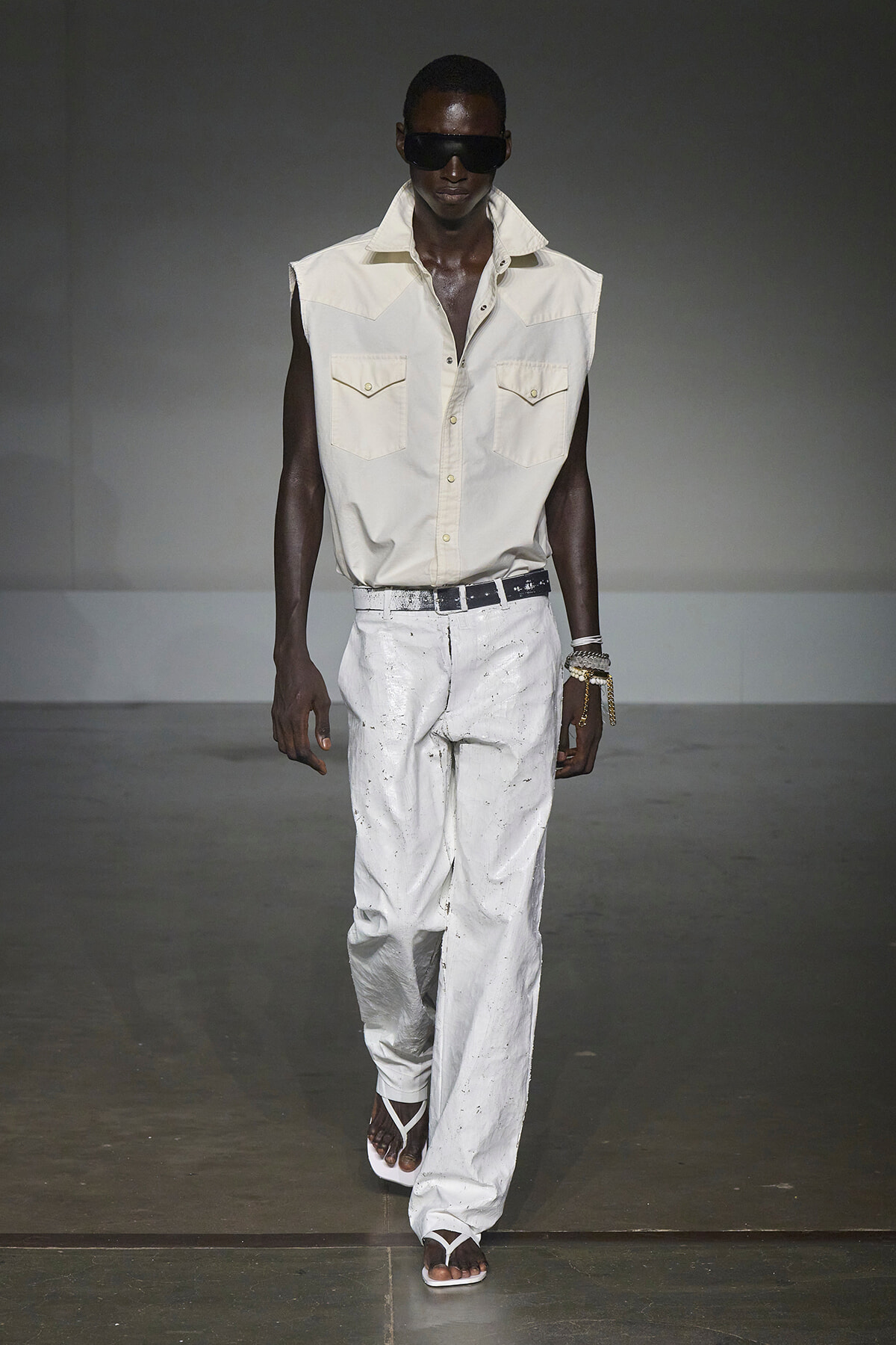 Male model on a runway wearing a sleeveless beige shirt with two breast pockets, white paint-splattered pants, a white belt, dark sunglasses, and white sandals.