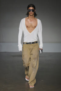 Male model on a runway wearing a deep V-neck white ribbed sweater, oversized sleeves, and beige paint-splattered wide pants with a black belt, sandals, and large sunglasses holding a small metallic clutch.