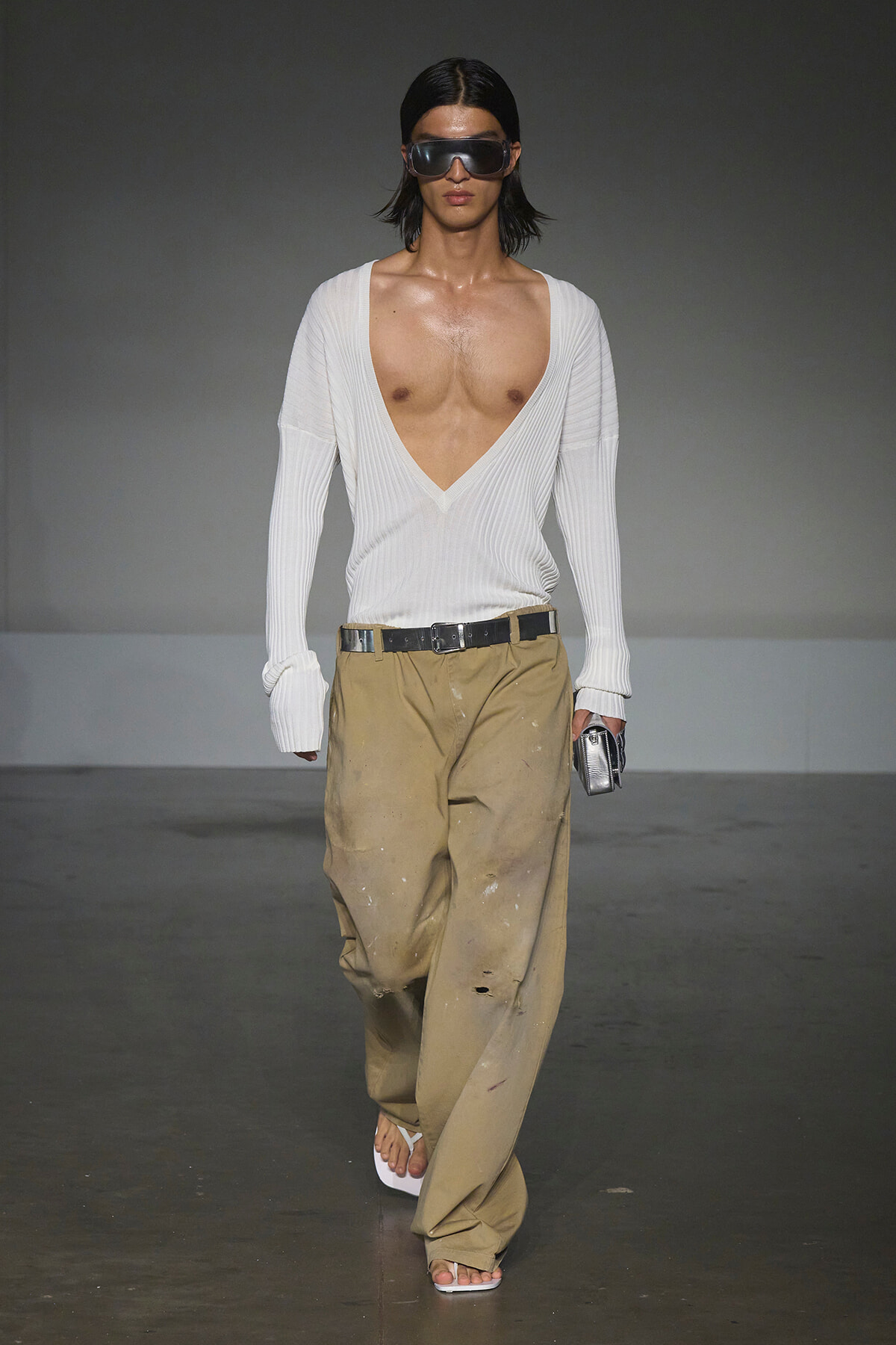 Male model on a runway wearing a deep V-neck white ribbed sweater, oversized sleeves, and beige paint-splattered wide pants with a black belt, sandals, and large sunglasses holding a small metallic clutch.