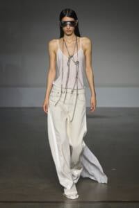 Model walks a runway in a pale pink satin camisole, oversized white wide-leg pants with side panels, and layered silver chains with futuristic sunglasses.