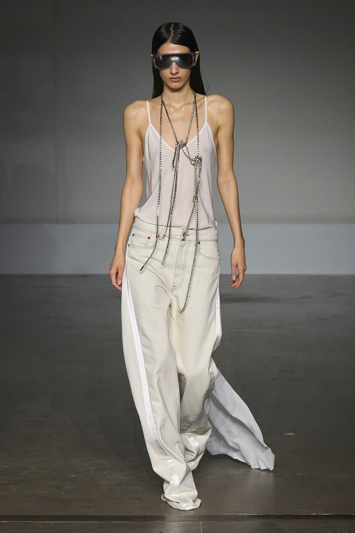 Model walks a runway in a pale pink satin camisole, oversized white wide-leg pants with side panels, and layered silver chains with futuristic sunglasses.