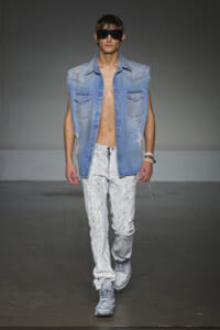Male model on a runway wearing a sleeveless light denim shirt, distressed paint-splattered jeans, gray sneakers, and large dark sunglasses.