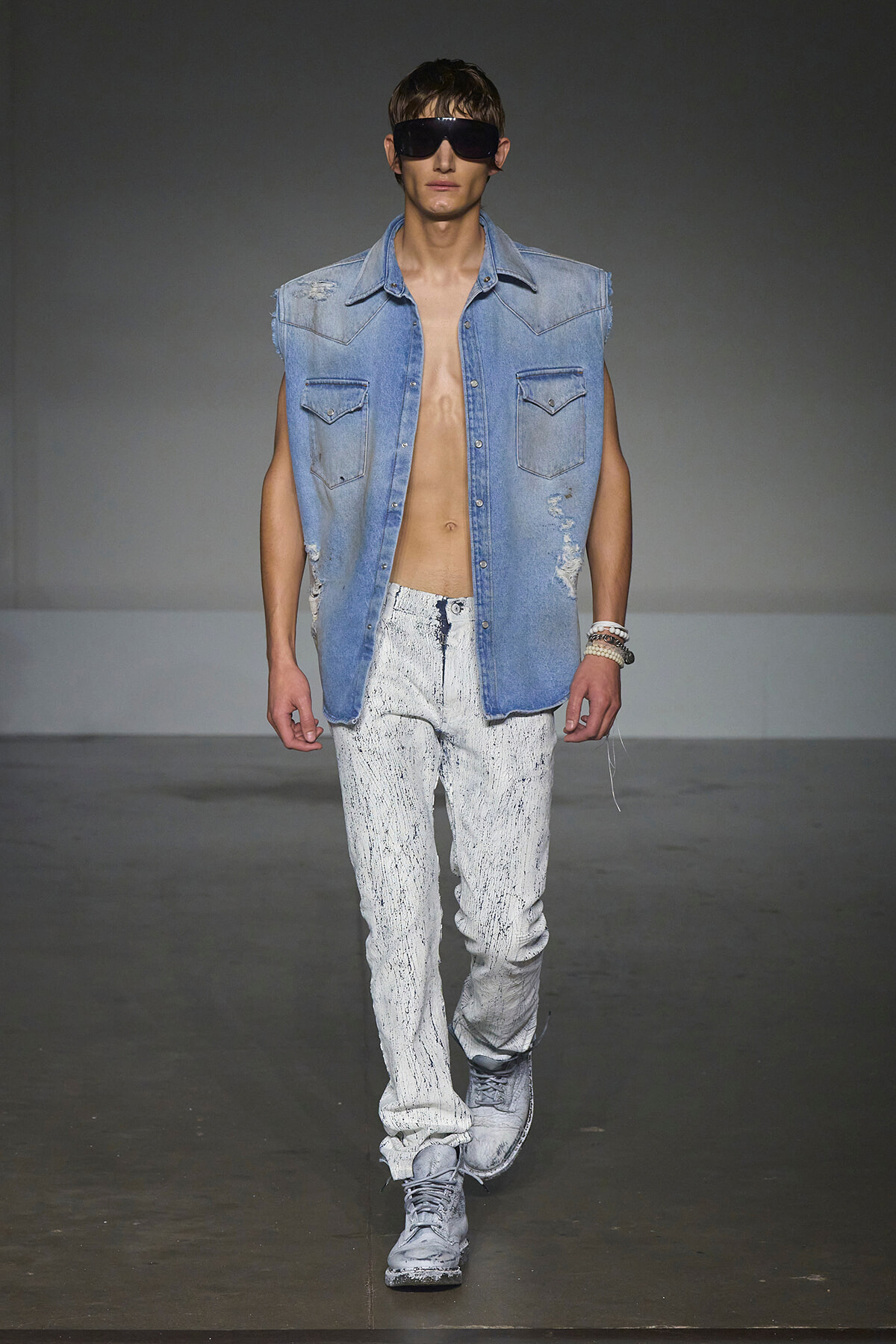 Male model on a runway wearing a sleeveless light denim shirt, distressed paint-splattered jeans, gray sneakers, and large dark sunglasses.