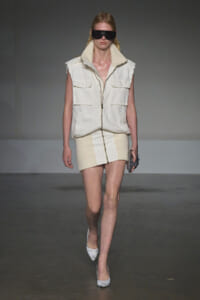 Model on a runway wearing a cream sleeveless zip vest with large pockets over a matching knit mini skirt, plus oversized black visor sunglasses and pointed heels.