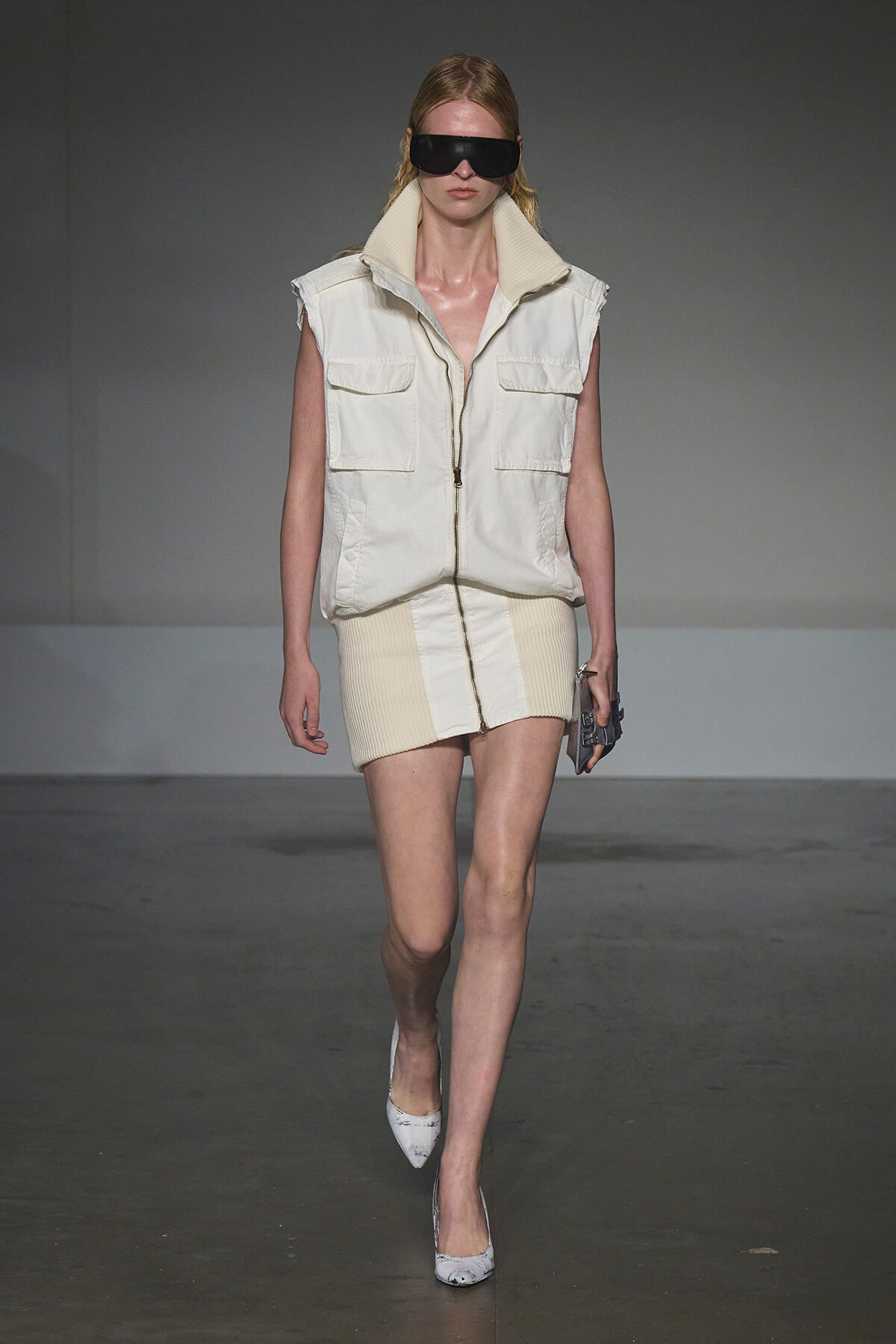 Model on a runway wearing a cream sleeveless zip vest with large pockets over a matching knit mini skirt, plus oversized black visor sunglasses and pointed heels.