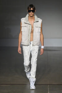 Male model walking runway in a sleeveless beige utility vest, white paint-splattered jeans, and white boots, wearing oversized black sunglasses.