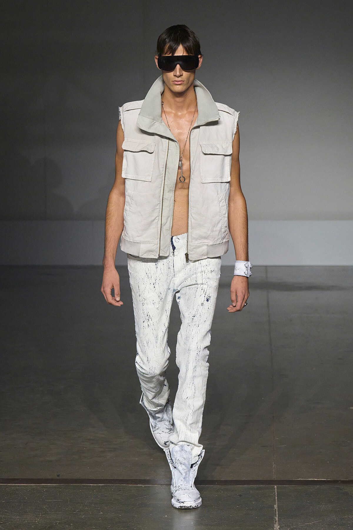 Male model walking runway in a sleeveless beige utility vest, white paint-splattered jeans, and white boots, wearing oversized black sunglasses.