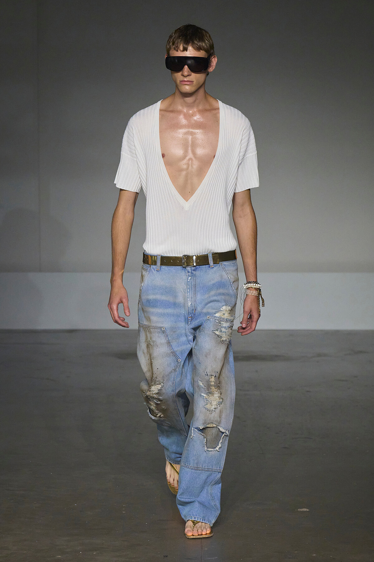 Male model walking a runway in a white deep-V ribbed top, oversized distressed jeans, brown belt, sandals, and large dark sunglasses.