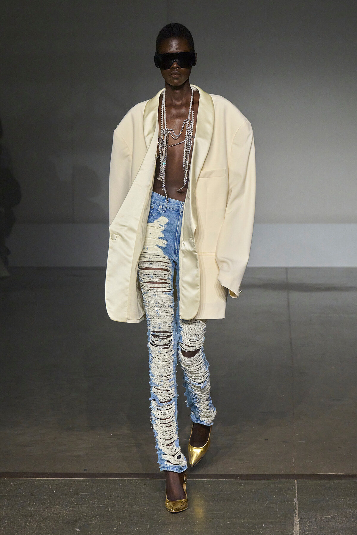 Model walks a fashion runway in an oversized cream blazer, heavily distressed light-wash jeans, gold pointed-toe heels, large dark sunglasses, and multiple layered pearl necklaces.