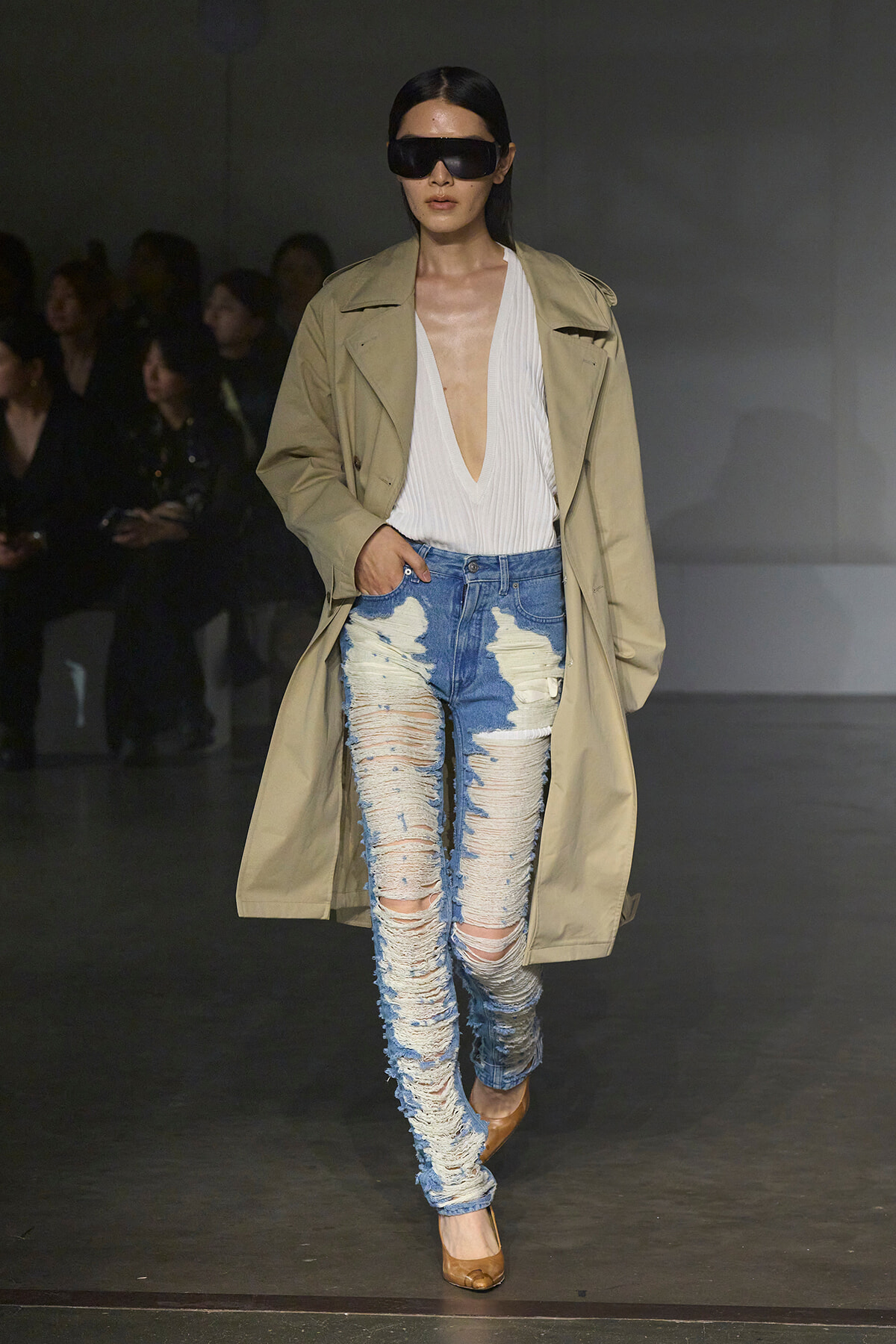 Model on a runway wearing a beige trench over a white plunging top, heavily ripped blue jeans, tan pumps, and oversized black sunglasses.