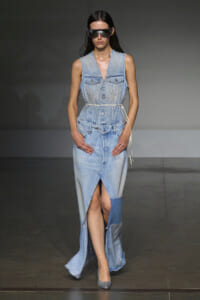 Model walks the runway in a light blue sleeveless denim dress with a thin belt, oversized sunglasses, and silver heels.
