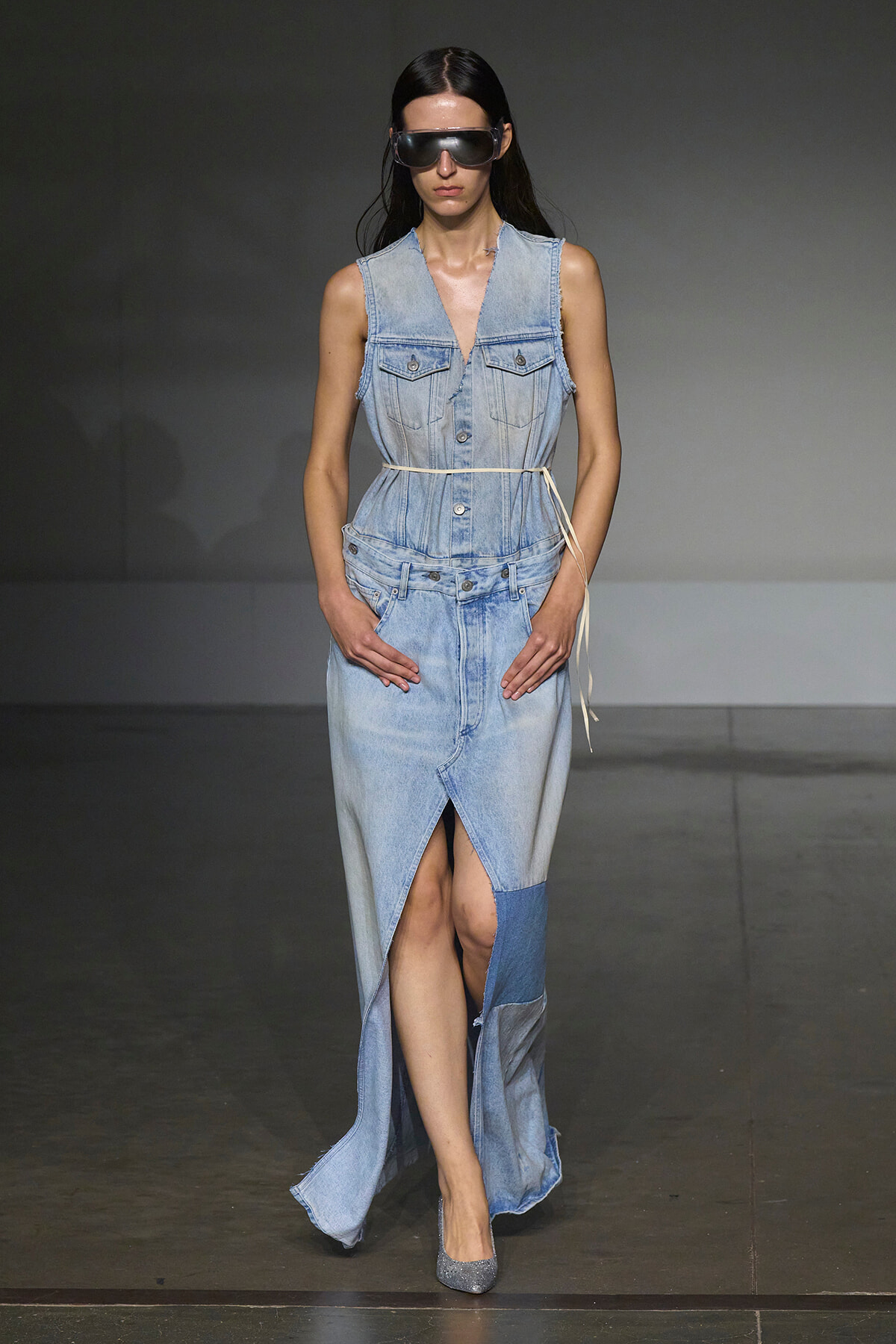 Model walks the runway in a light blue sleeveless denim dress with a thin belt, oversized sunglasses, and silver heels.