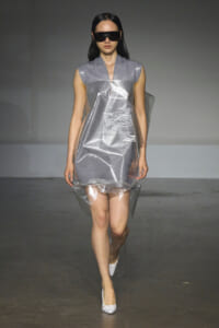 Model walks a runway in a futuristic metallic silver dress with a clear plastic overlay, oversized black sunglasses, and white heels.