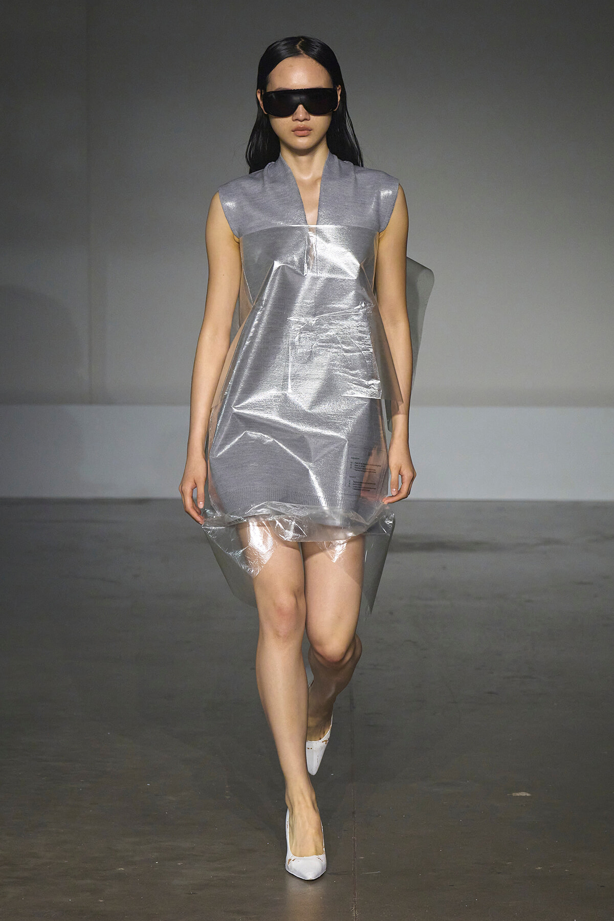 Model walks a runway in a futuristic metallic silver dress with a clear plastic overlay, oversized black sunglasses, and white heels.