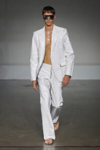 Male model walks the runway in a white oversized suit with no shirt, wearing large futuristic sunglasses.
