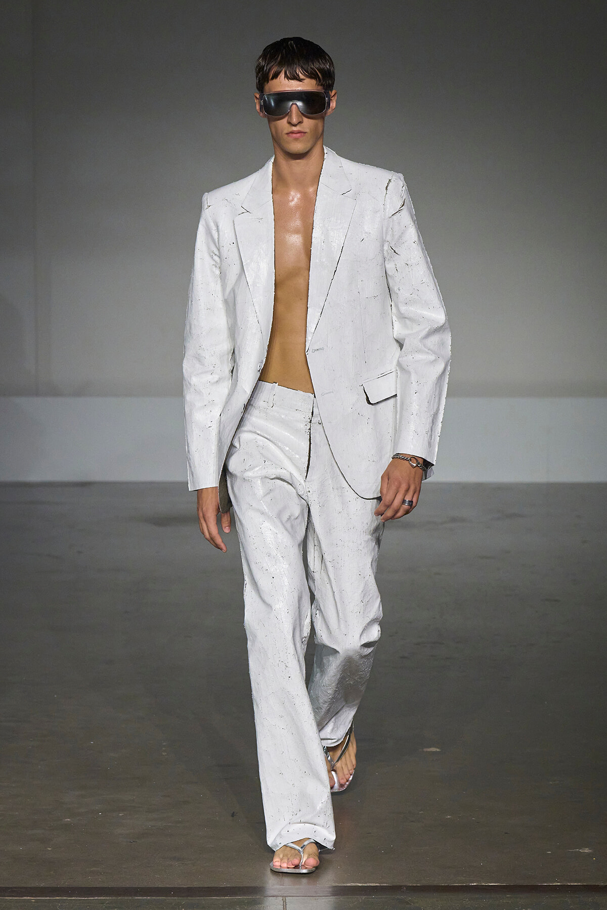 Male model walks the runway in a white oversized suit with no shirt, wearing large futuristic sunglasses.