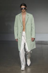 Male model on a runway wearing a pale mint-green long coat over bare chest and white metallic pants with oversized sunglasses.