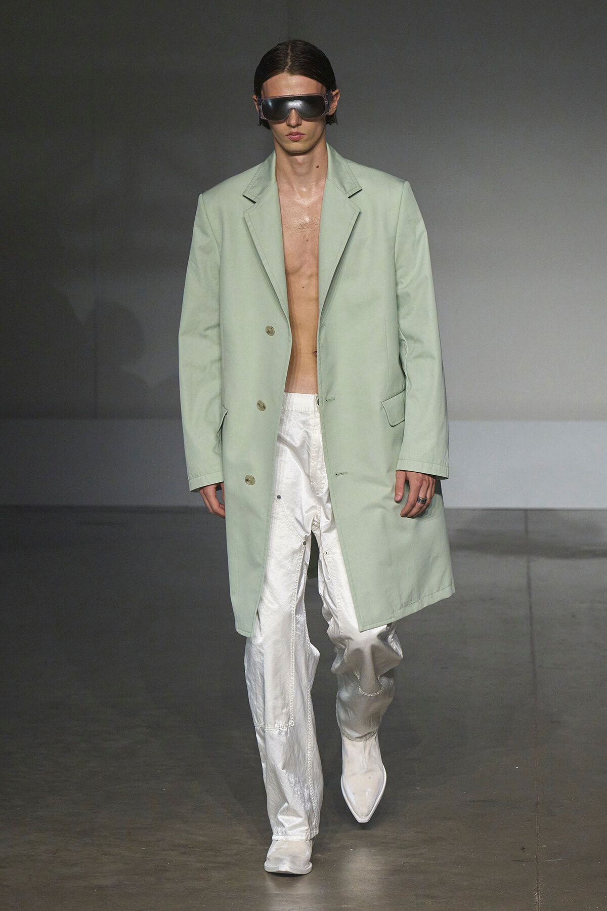 Male model on a runway wearing a pale mint-green long coat over bare chest and white metallic pants with oversized sunglasses.
