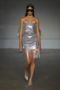 Model walking runway in a metallic strapless dress with a shiny, crinkled texture and silver glitter heels, wearing large black futuristic sunglasses.