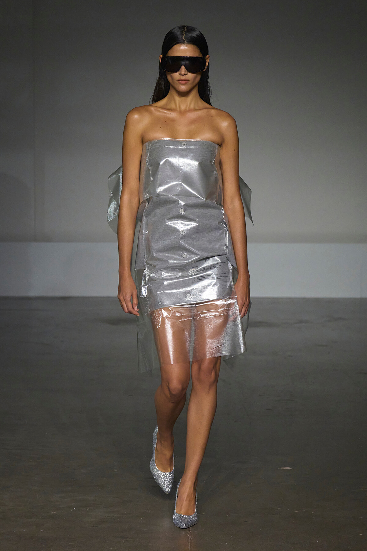 Model walking runway in a metallic strapless dress with a shiny, crinkled texture and silver glitter heels, wearing large black futuristic sunglasses.