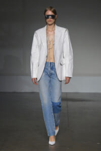 Runway model wearing an oversized white blazer, light denim jeans, white heels, oversized sunglasses, and long layered chains.
