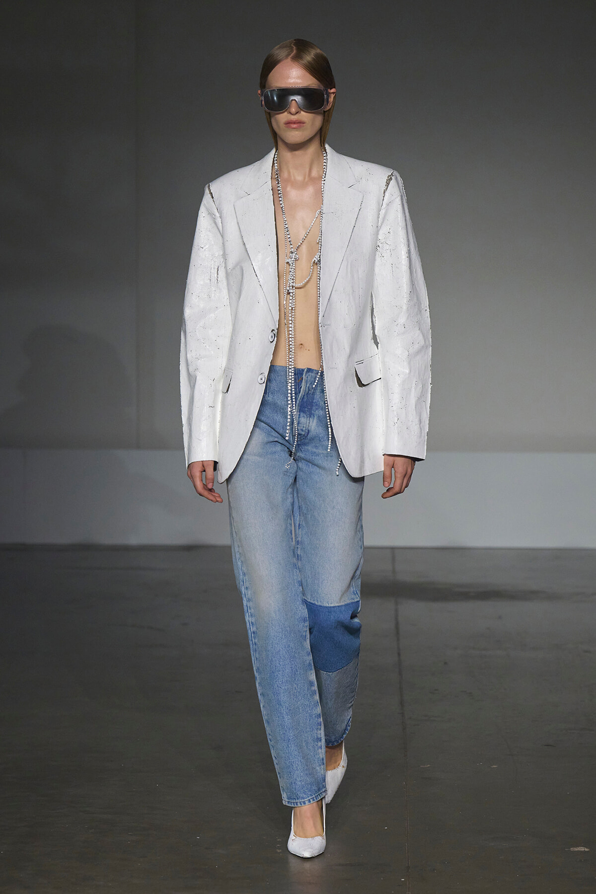 Runway model wearing an oversized white blazer, light denim jeans, white heels, oversized sunglasses, and long layered chains.