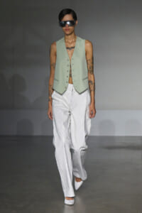 Model walking a runway in a mint sleeveless vest with a button front over white wide-leg pants and oversized visor sunglasses.