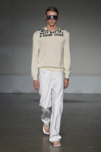 Male model walking the runway in a cream knit sweater with black text around the neckline, paired with white wide-leg pants and white sandals, wearing dark sunglasses.