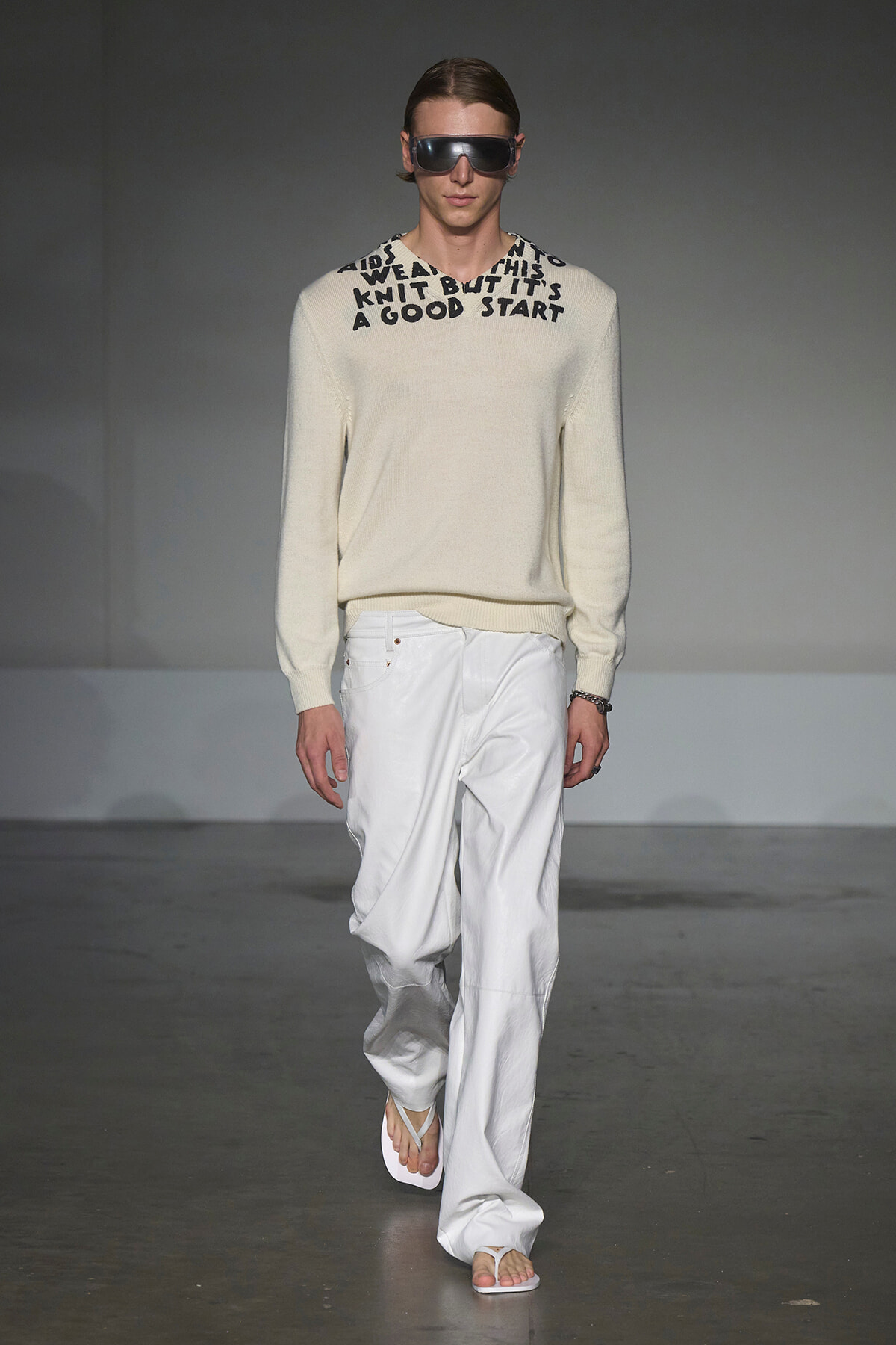 Male model walking the runway in a cream knit sweater with black text around the neckline, paired with white wide-leg pants and white sandals, wearing dark sunglasses.