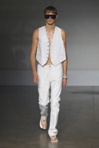 Male model on a runway wearing a sleeveless white vest, white jeans, white flip-flops, and oversized black sunglasses.