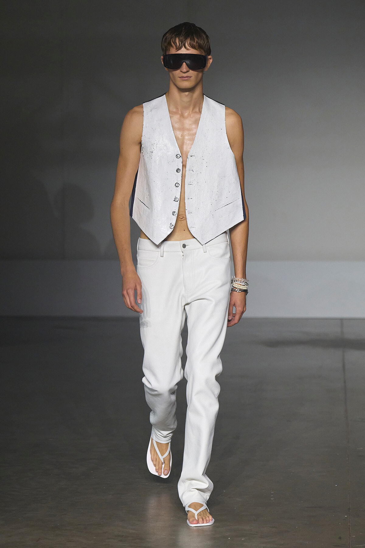 Male model on a runway wearing a sleeveless white vest, white jeans, white flip-flops, and oversized black sunglasses.