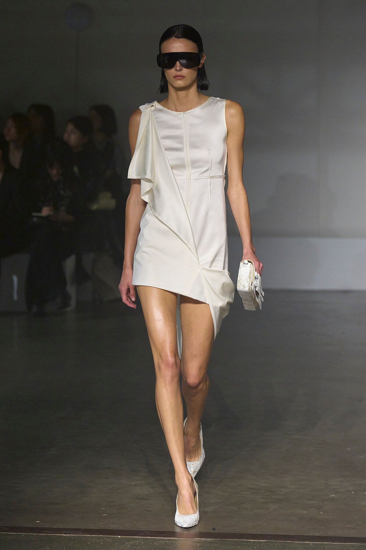 Model walks runway in a sleeveless white dress with a draped asymmetric overlay and matching glittery heels, holding a small clutch and wearing oversized sunglasses.