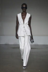 Runway model in a white sleeveless blazer and matching wide-leg pants, wearing oversized sunglasses and holding a silver glitter clutch.