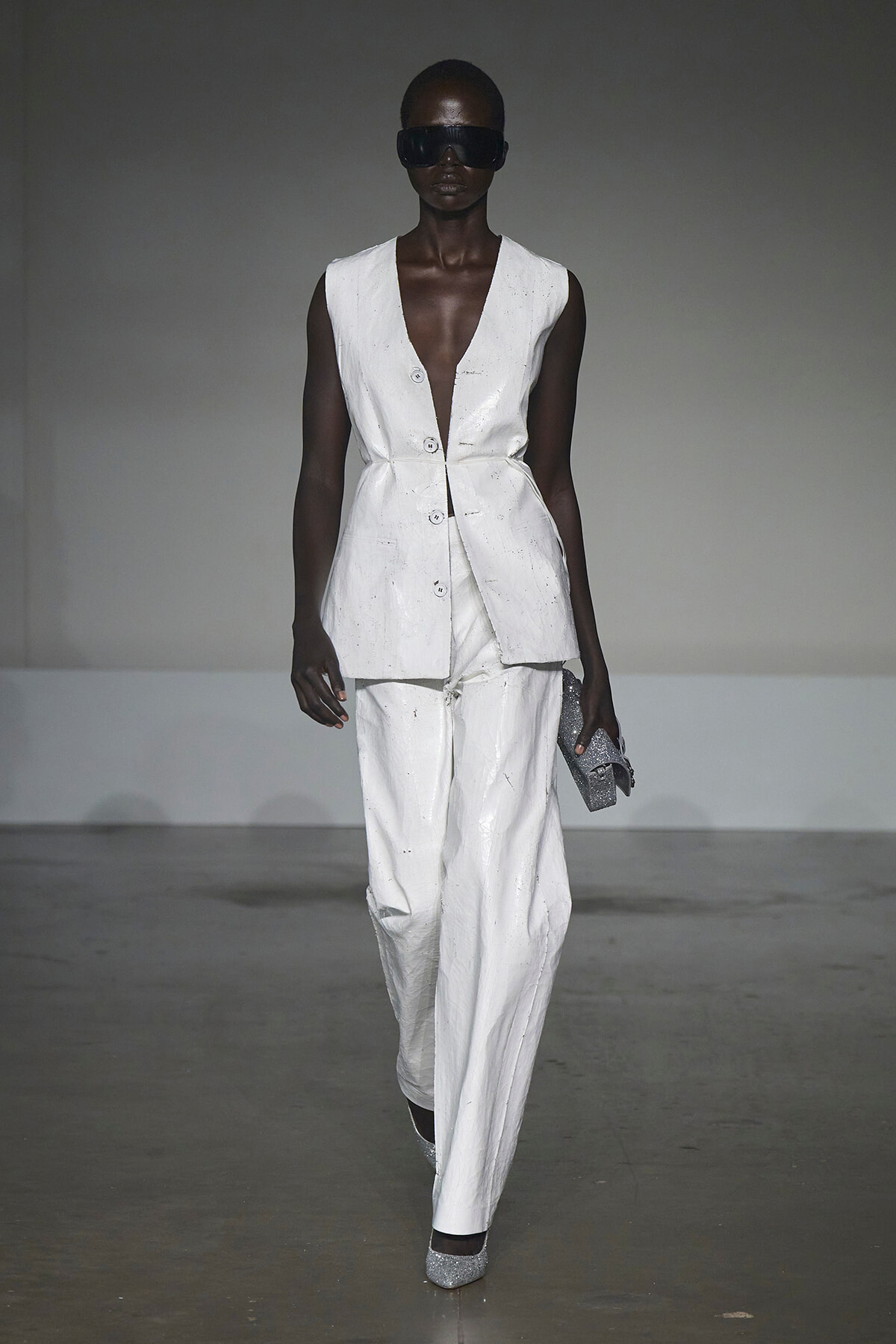 Runway model in a white sleeveless blazer and matching wide-leg pants, wearing oversized sunglasses and holding a silver glitter clutch.
