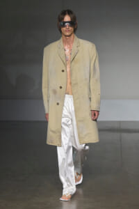 Male model walks the runway in a beige long coat over white satin pants, wearing oversized sunglasses and flip-flop sandals, with visible chest tattoos and a gray backdrop.