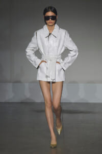 Model walking the runway in a white belted trench coat with large collar and black sunglasses, gold glitter heels.