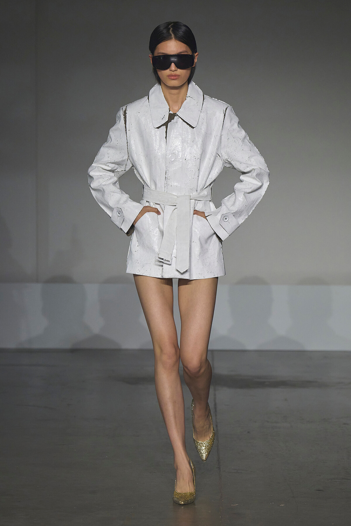 Model walking the runway in a white belted trench coat with large collar and black sunglasses, gold glitter heels.