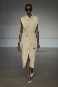 Runway model wearing a sleeveless beige belted trench dress with a front slit, silver glitter heels, and oversized black sunglasses.