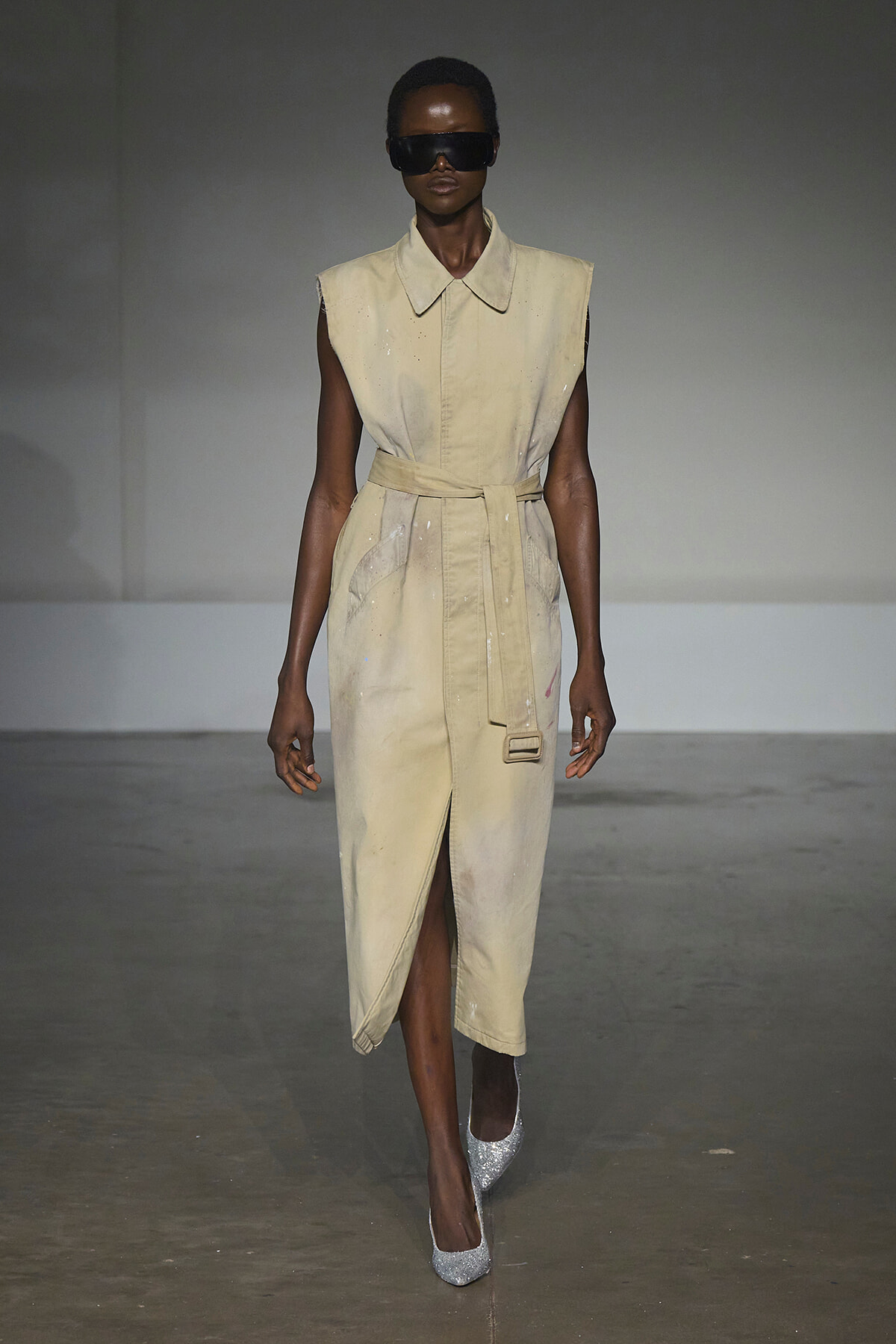 Runway model wearing a sleeveless beige belted trench dress with a front slit, silver glitter heels, and oversized black sunglasses.