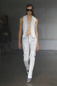 Model walking runway in a sleeveless white vest over distressed pale jeans with pearl chains draped across the torso and oversized black sunglasses, silver ankle boots visible.